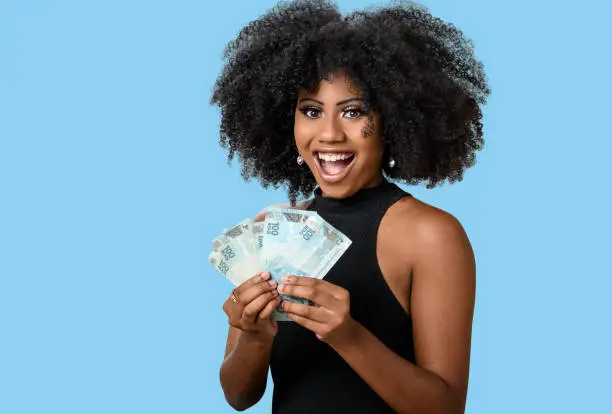 Photo of young black woman smiling holding brazilian money bills, positively surprised, space for text, person, advertising concept, isolated on blue background.