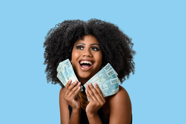 young black woman smiling holding brazilian money bills, positively surprised, space for text, person, advertising concept, isolated on blue background. young black woman smiling holding brazilian money bills, positively surprised, space for text, person, advertising concept, isolated on blue background. brazilian currency photos stock pictures, royalty-free photos & images