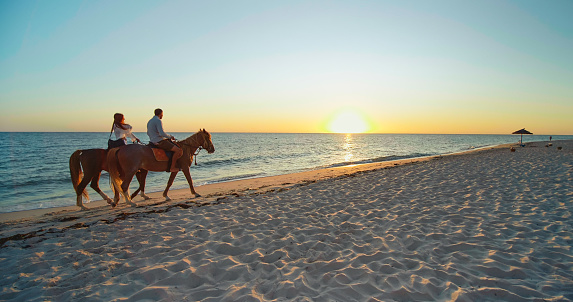 Monastir, Tunisia, 2022: Couple walking at sunset, with horses on a beach. Beautiful view with horseback riding, love, holiday, destinations, seaside, holiday activities. Powerful horses on the beach.
