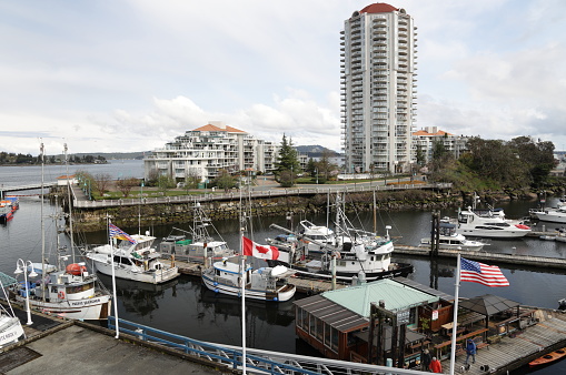 Nanaimo, Canada - April 10, 2022: Flags line Harbourfront Walkway by the marina in the Port of Nanaimo. The promenade is a popular viewing area near commercial and recreational vessels. Upscale residential buildings stand in the background. Spring afternoon with cloudy skies.