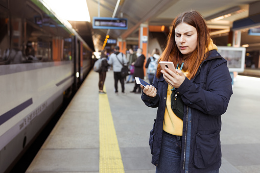Young redhhead woman waiting on station platform with backpack on background electric train using smart phone. Railroad transport concept