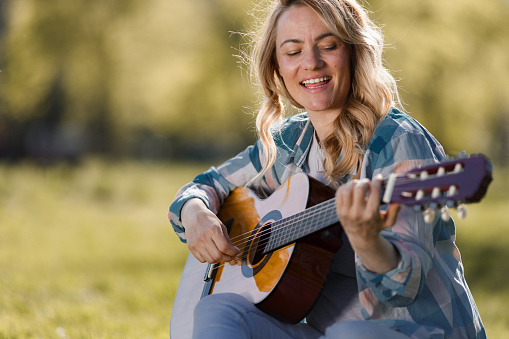 Happy woman sitting on the maedow playing guitar and singing during spring day