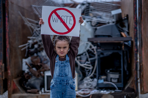 A young girl of Hawaiian decent stands in front of a garbage depot as she holds up a sign that reads 