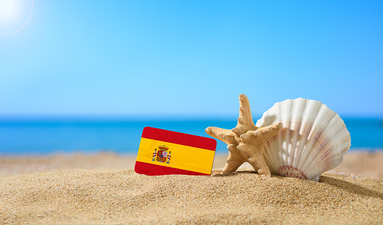 Tropical beach with seashells and Spain flag. The concept of a paradise vacation on the beaches of Spain.
