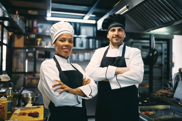 Portrait of confident professional cooks looking at camera. Male chef and his African American female coworker standing with arms crossed and looking at camera. chefs whites stock pictures, royalty-free photos & images