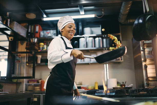 happy black female chef preparing food in frying pan at restaurant kitchen. - restaurant imagens e fotografias de stock