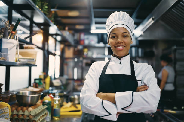 Portrait of confident black female chef at restaurant kitchen looking at camera. Happy African American woman standing with arms crossed while working as chef in a restaurant and looking at camera. chefs whites stock pictures, royalty-free photos & images