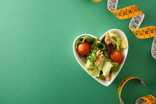 Concept of diet and health with heart-shaped bowl with salad with nuts and metric tape on a green background. Top view. Horizontal composition.