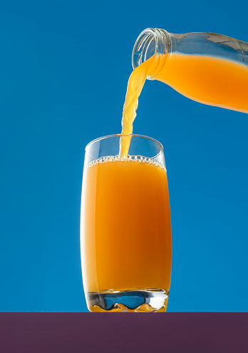 Pouring orange juice from a bottle into a glass, isolated on a blue background. Low angle view with a glass of fresh orange juice.