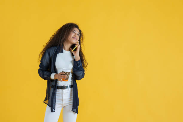 Young woman talking on phone while holding coffee cup on yellow background stock photo