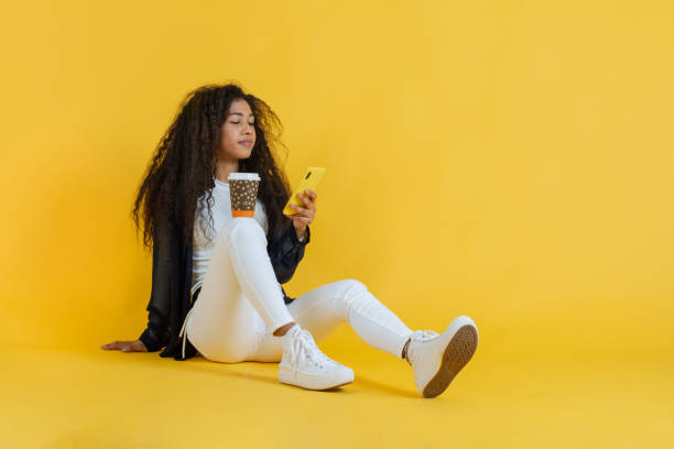 Young woman sitting on floor using phone with cup of coffee on her knee stock photo
