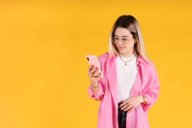 mujer joven de pie usando su teléfono celular sobre fondo amarillo - vertical studio shot indoors pink fotografías e imágenes de stock