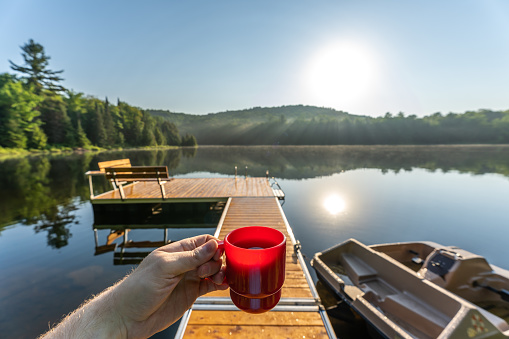 POV photo of a man having a coffee by a small and quiet fishing lake in the Laurentides (Laurentians), Quebec, during a sunrise of summer.