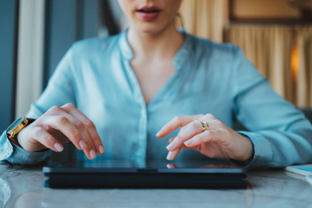 Anonymous woman sitting in the office. The tablet is laid down on the table and she is using it. She is wearing a light blue blouse.