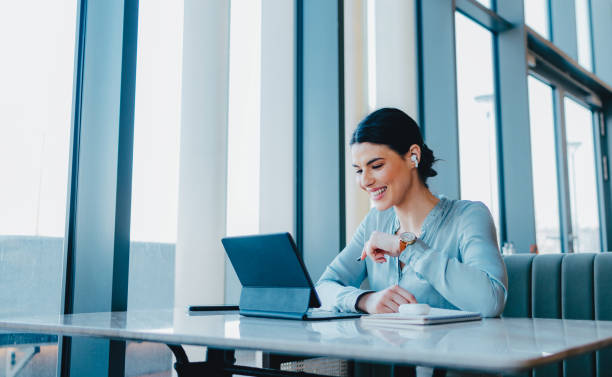 Beautiful Woman Wearing Earbuds And Watching Something Fun on Her Tablet Beautiful happy woman with dark hair, sitting at her desk. She is wearing earbuds and looking down at her tablet. There is a earbuds case and a notebook on the desk. remote location stock pictures, royalty-free photos & images