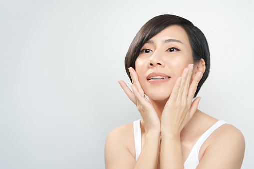 Studio portrait of Asian short hair middle aged woman wear white tank top, put hands on her cheeks and smile on white isolated background.