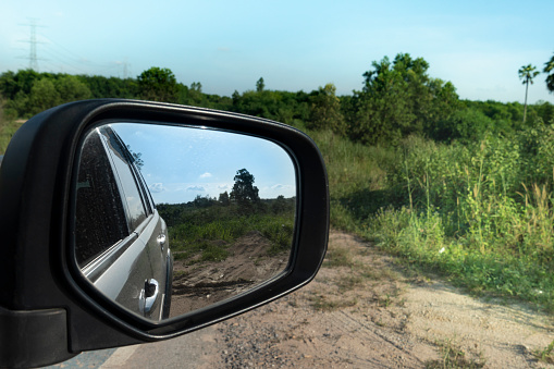 Abstract view of nature beside cars from behind through the mirrors wing of a gray car. Beside of road with soil and green grass and trees under blue sky.