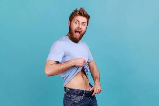 Portrait of bearded man having willpower points at slim waist in big trousers, successful weight loss, keeping diet and going for sport. Indoor studio shot isolated on blue background.