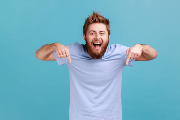 Man pointing down and looking at camera showing place for presentation, expressing positive emotions Portrait of happy satisfied bearded man pointing down and looking at camera, showing place for idea presentation, expressing positive emotions. Indoor studio shot isolated on blue background. aiming stock pictures, royalty-free photos & images