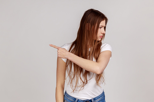 Portrait of resentful upset attractive woman turning away with disgust and ordering to leave, showing way out, feeling betrayed, wearing white T-shirt. Indoor studio shot isolated on gray background.