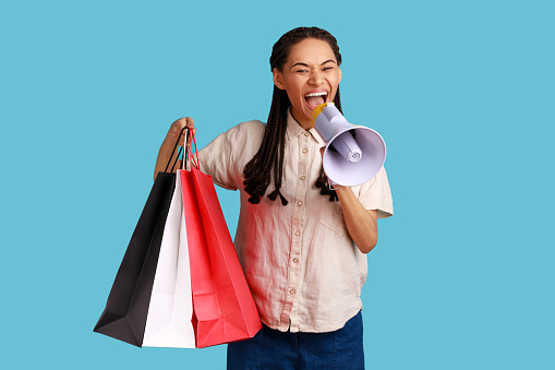 Portrait of excited woman with black dreadlocks screaming in megaphone and holding shopping bags, advertising discounts, wearing white shirt. Indoor studio shot isolated on blue background.