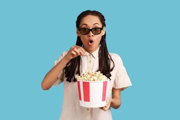 Excited woman with dreadlocks in 3d imax glasses watching movie film, hold popcorn, watching interesting movie, keeping mouth open, wearing white shirt. Indoor studio shot isolated on blue background.