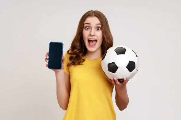 Photo of Portrait of excited female of young age looking at camera with surprised expression and open mouth, holding soccer ball and cell phone with blank screen.