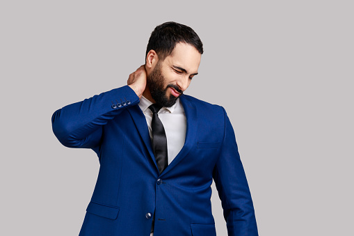 Bearded businessman touching neck feeling pain and numbness, worried about muscle tension, osteochondrosis, wearing official style suit. Indoor studio shot isolated on gray background.