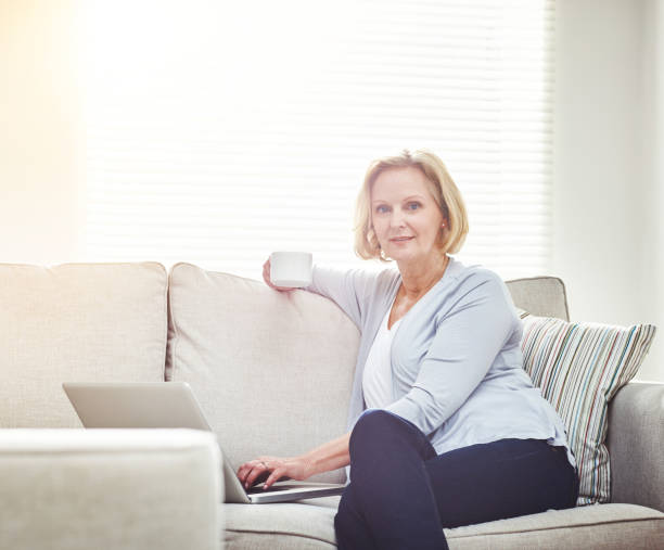 istock Mature woman drinking coffee and browsing online on a laptop while relaxing alone on a sofa at home 1394125303