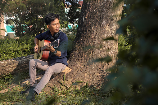 Portrait of relaxing Asian man in casual shirt playing guitar at the park, leaning against the tree