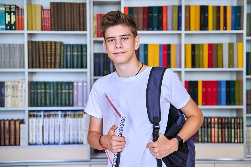 Portrait of male teenage student looking into the camera in library. Smiling positive guy 16, 17 years old with backpack, notebooks. Education, high school, college, adolescence concept