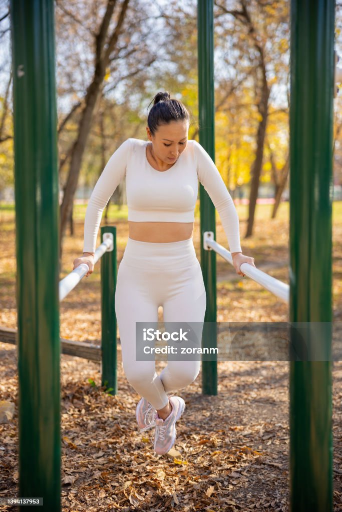 Asian woman in casual sports clothes working out on parallel bars in public park Asian woman in casual sports outfit exercising arm muscles on parallel bars in the park, bending legs in knee, leisure outdoor workout on sunny autumn day 35-39 Years Stock Photo