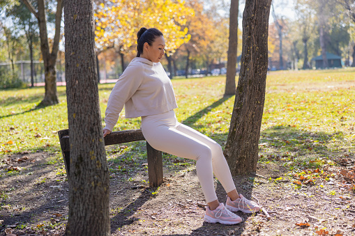 Asian woman in casual sports clothes exercising muscles and doing dips, leaning on log hurdle in the park and looking down, outdoor activities on sunny autumn day