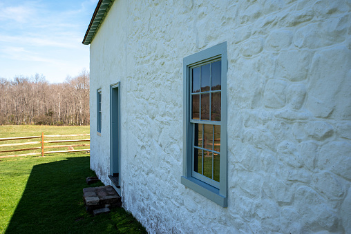 Beautiful sude view of a whitewashed stone cottage with blue window rim and antique window panes with rural scene reflection. Stone texture and green pasture and woodland background in late afternoon natural light with copy space and no people.
