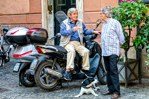 Rome, Italy, June 04 -- Two Italian adults converse animatedly in a scene of daily life along via dei Coronari, near Piazza Navona, in the historic and baroque heart of the Eternal City. Called Via Recta in medieval times, Via dei Coronari is still today one of the main pedestrian routes to St. Peter's Basilica from the center of Rome and from the Piazza Navona area. The Rione Parione, the district surrounding Piazza Navona, is one of the most beautiful and visited districts of Rome for the presence of countless artistic and historical treasures, monuments and ancient Romanesque and Baroque churches, but also for its squares and hidden alleys to explore freely, where it is easy to find typical restaurants, small artisan shops, street artists, and the original Roman soul. In 1980 the historic center of Rome was declared a World Heritage Site by Unesco. Image in high definition format.