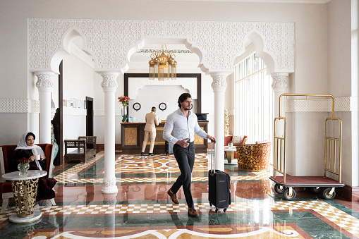 Full length view of Middle Eastern man in casual business attire rolling wheeled luggage across elegant marble floor after checking out.