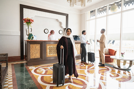 Full length view of smiling Middle Eastern woman in traditional attire walking through sunny reception area with male guests talking in background.
