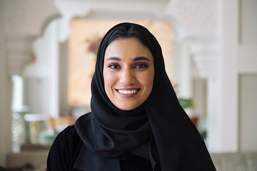 Indoor portrait of woman in traditional black abaya and hijab smiling at camera while standing in hotel lobby with Islamic architectural design.