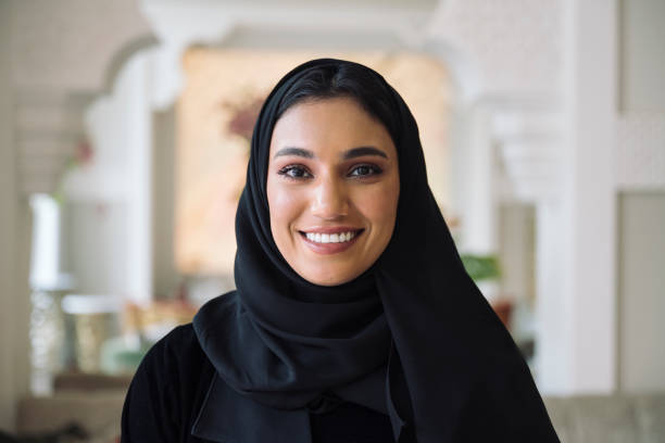 Headshot of early 20s Middle Eastern woman Indoor portrait of woman in traditional black abaya and hijab smiling at camera while standing in hotel lobby with Islamic architectural design. middle eastern culture stock pictures, royalty-free photos & images
