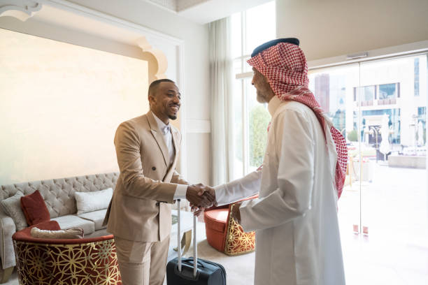 Middle Eastern and Black businessmen shaking hands in hotel Mature Saudi man in dish dash, kaffiyeh, and agal welcoming smiling associate in western dress as he arrives for business conference in Riyadh. global business handshake stock pictures, royalty-free photos & images