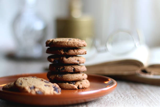 Stack of chocolate chip cookies, open book and reading glasses, lit candle and vase with flowers on the table. Hygge at home. Selective focus.