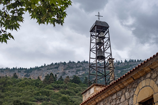 Greek culture and orthodox church architecture. Place of worship for Christianity. Background with copy space. Beautiful metal made bell tower with three bells against mountain view on grey cloudy day.