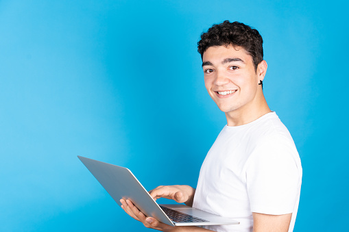 Portrait of hispanic teenager boy working on laptop and looking at camera isolated on blue background.