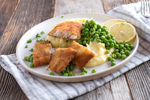 Delicious homemade traditional fish dish with breaded redfish fillet, mashed potatoes and buttered green peas on a plate isolated on wooden table background