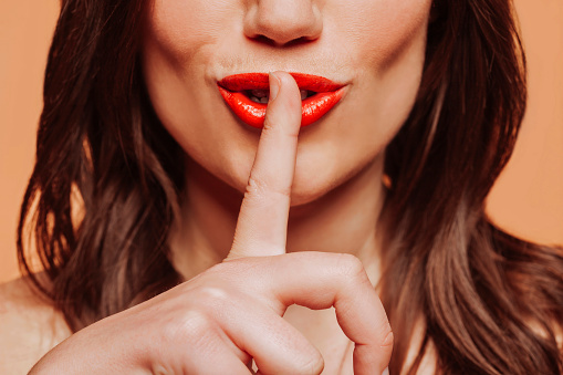 Close up portrait of young brunette woman with red lips doing silence gesture with finger on mouth. Shh! Shut up.\