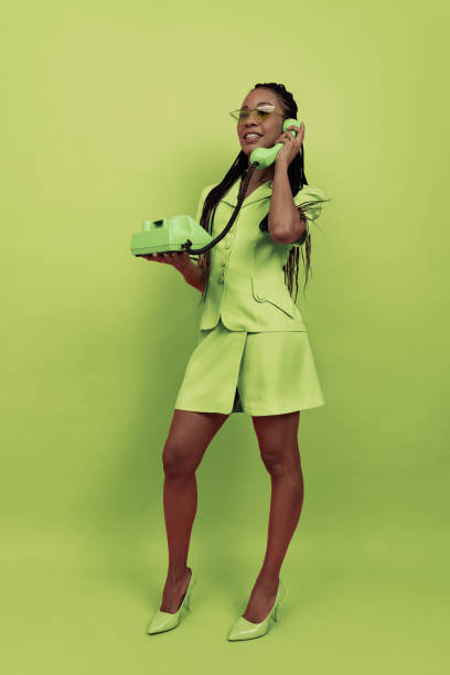 monochromatyczny portret uśmiechniętej afrykańskiej dziewczyny w stroju w stylu retro trzymającej zabytkowy telefon odizolowany na zielonym tle. pojęcie emocji, piękna, sztuki, mody, młodości, stylu - fashion youth culture women old fashioned zdjęcia i obrazy z banku zdjęć