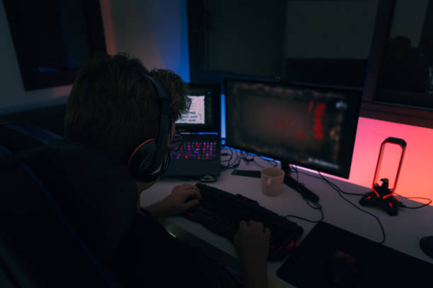 istock Young blond guy playing video games on a computer with headphones and using a backlit coloured keyboard. 1394180510