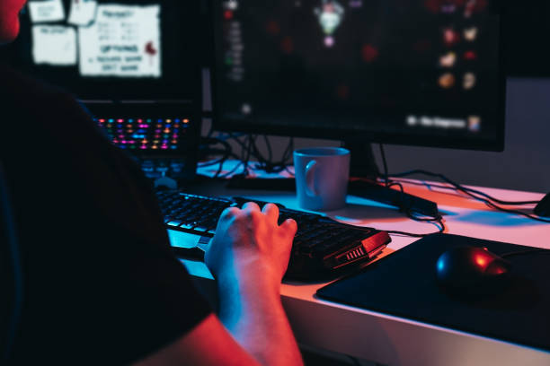 Close-up of the hands of a young online video game professional, playing on the computer. Close-up of a gamer set-up Close up of a gamer montage, detail of the hands of a young man with short blond hair, wearing glasses and headphones, dressed in black shirt, addicted to online video games, playing. Young gamer illuminated with blue and red lights. home office. Dim light from computer monitor, dark room, coloured lights. Horizontal. gaming chair photos stock pictures, royalty-free photos & images