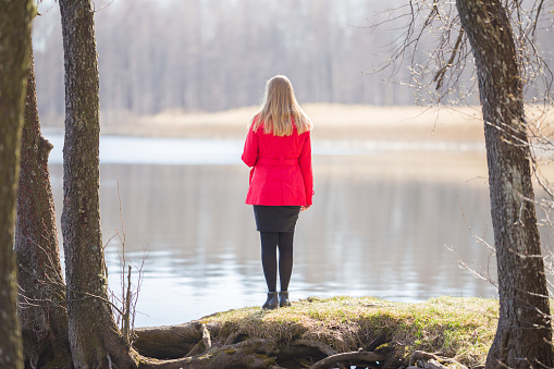 Young adult woman in red coat standing at lake shore between trees and looking far away. Thinking about life. Peaceful atmosphere in sunny spring morning. Spending time alone in nature. Back view.