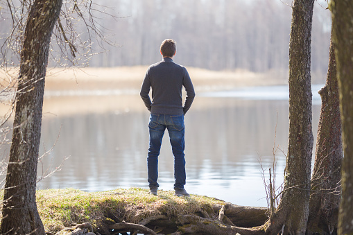 Young adult man in dark clothes standing at lake shore between trees and looking far away. Thinking about life. Peaceful atmosphere in sunny spring morning. Spending time alone in nature. Back view.